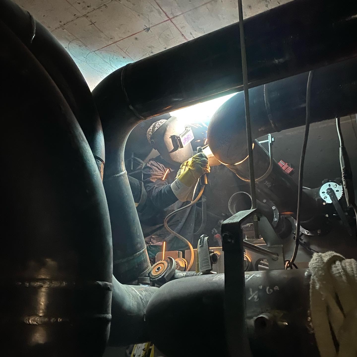 Welder in a hood welding large black pipe in a tight mechanical space