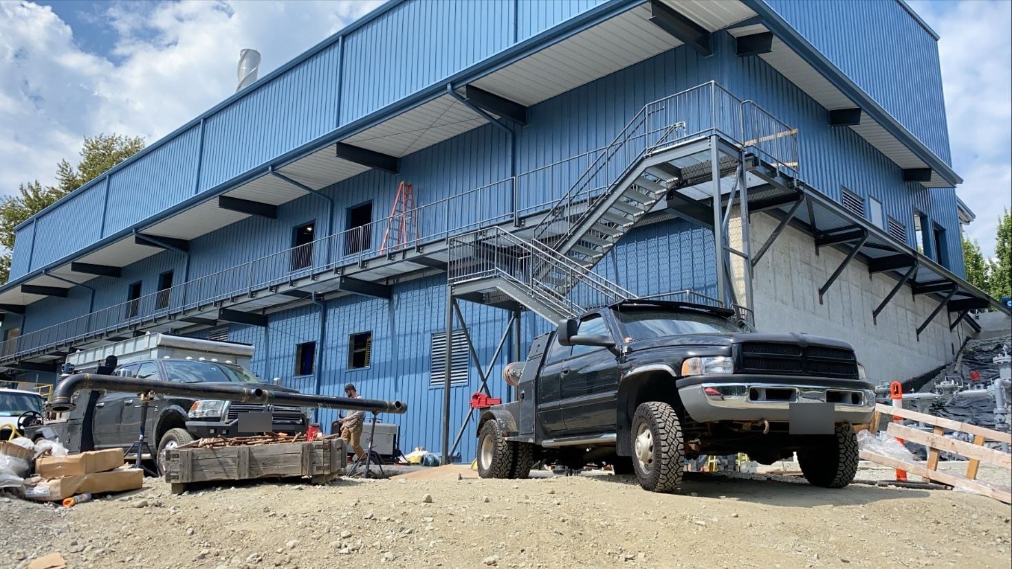 Berto's welding truck parked below the steel stairs of a blue industrial building, pipe on stands nearby