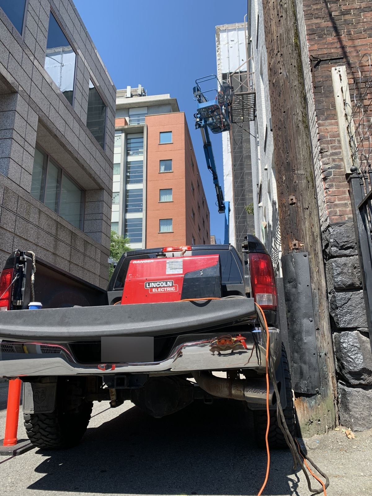 Pickup with a Lincoln Electric engine-driven welder backed into a narrow city laneway, a boom lift working on the building above