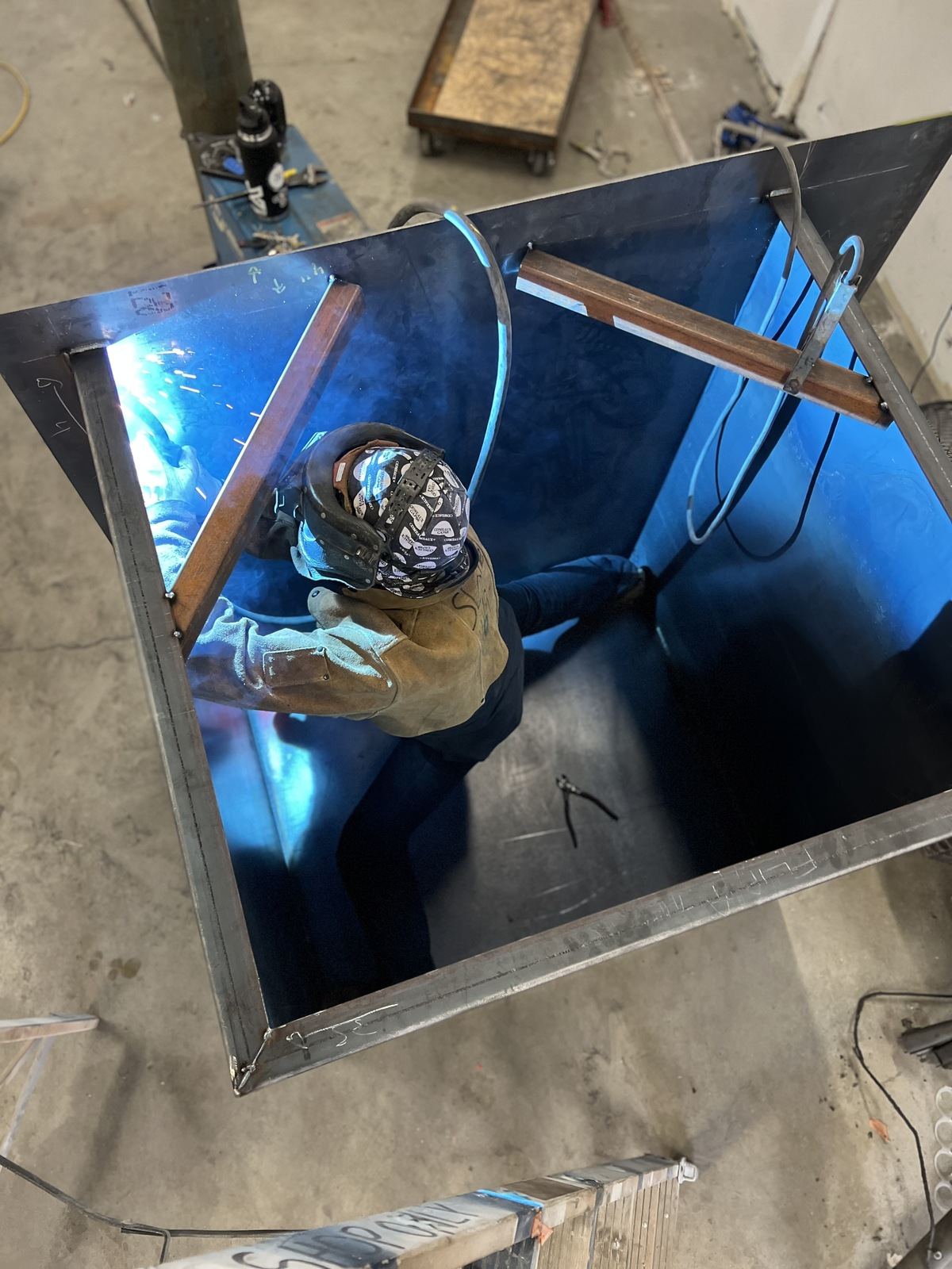 Welder in a helmet working inside a steel enclosure, lit by the welding arc