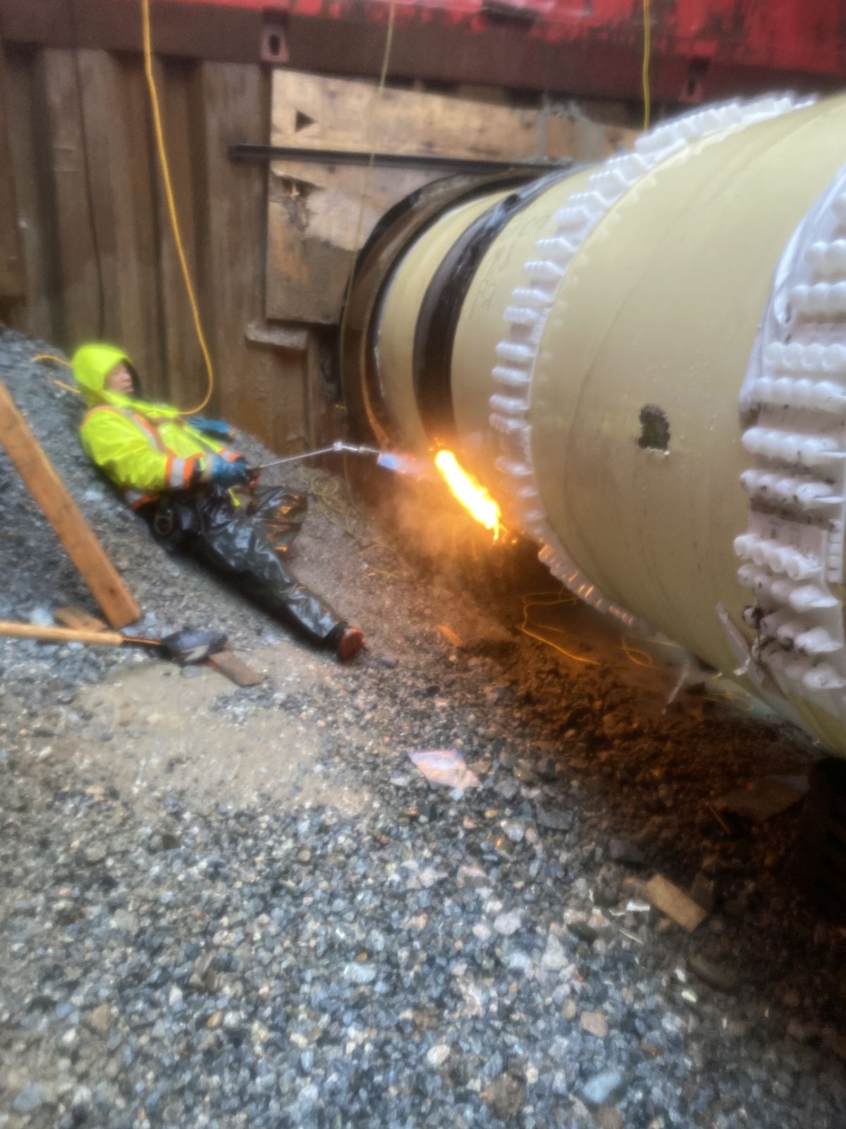 Welder in hi-vis welding a large-diameter pipe joint in a gravel trench, sparks flying