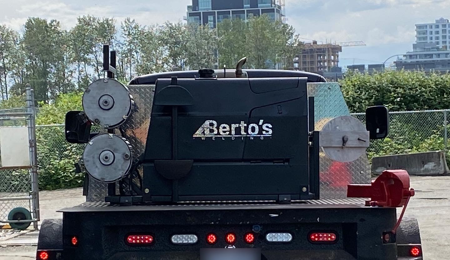 Rear view of the Berto's mobile welding truck with its branded welder and cable reels, city towers behind