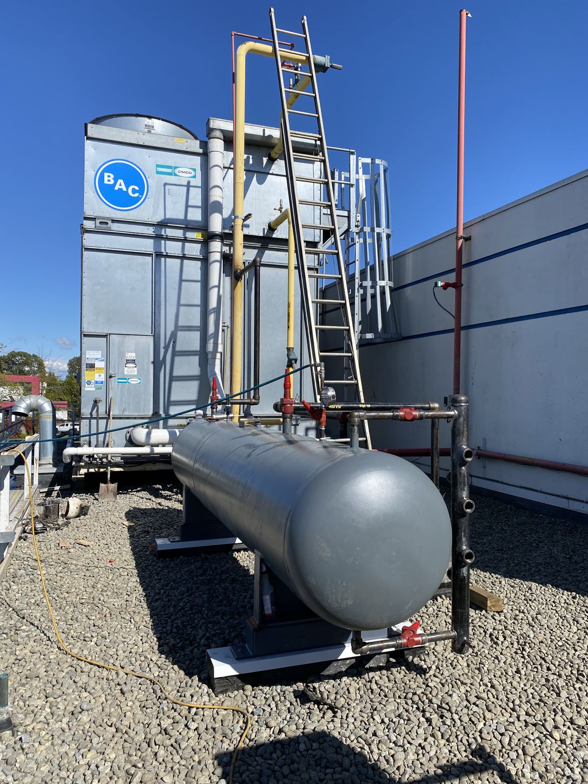 Horizontal steel tank with piping on a gravel rooftop pad beside a cooling tower